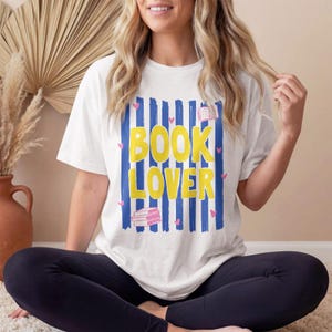 Peut inclure: Un t-shirt blanc à manches courtes avec un motif graphique composé de rayures verticales bleues, du texte jaune "BOOK LOVER", de petits cœurs roses et des illustrations de livres empilés. Le t-shirt a un col rond et une coupe décontractée.