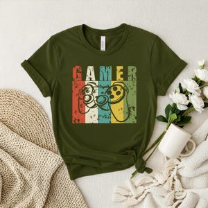 Peut inclure: T-shirt vert olive avec un design rétro. Le mot "GAMER" est au-dessus de deux manettes de jeu. Les manettes sont soulignées en noir avec des rayures rouges, blanches, bleues et jaunes derrière elles. Le t-shirt est en matière souple.