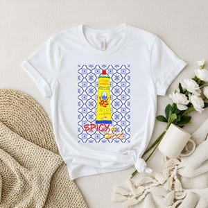 Peut inclure: T-shirt blanc avec un motif d'un tube jaune de pâte harissa, des accents rouges et bleus, et un motif de carreaux bleus et blancs. Le mot "SPICY" est imprimé en rouge. Le tube a un bouchon rouge.