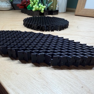 May include: Two black, geometric, honeycomb-shaped structures. One is oval, the other is rectangular. The structures are made of a series of intersecting, vertical, and horizontal lines, creating a grid-like pattern. They are placed on a light brown wooden surface.