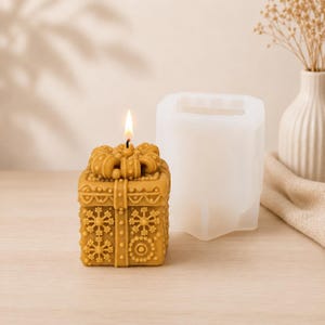 Peut inclure: Une bougie carrée jaune doré en forme de boîte cadeau avec un nœud sur le dessus, ornée de motifs de flocons de neige et de cercles en relief. Une mèche allumée brûle au centre. À côté se trouve un moule en silicone blanc translucide.