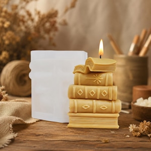 May include: A stack of book-shaped candles with a lit wick, next to a white mold. The candles are a warm yellow color, with detailed book spine designs. The background includes a wooden surface and neutral-toned decor.