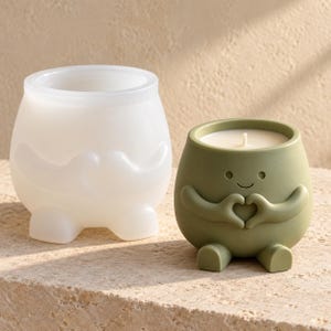 May include: A translucent white silicone mold shaped like a small character with arms forming a heart and a finished olive green candle in an identical container with a white wax surface and a centered wick, both featuring a smooth rounded base.