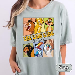 Pode incluir: Uma t-shirt verde sálvia claro com uma estampa gráfica de personagens de O Rei Leão. O design inclui Pumbaa, Simba, Timon, Scar, Zazu e Rafiki numa grelha com o texto THE LION KING EST. 1994 impresso em amarelo.