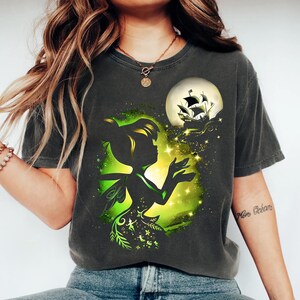 May include: A charcoal gray t-shirt featuring a graphic print of a fairy silhouette in front of a glowing green light, surrounded by sparkling stars and a pirate ship sailing toward a large full moon in the upper right corner of the design.