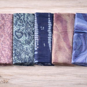 May include: An overhead shot displays five folded scarves in shades of pink, blue, and purple. The scarves feature various patterns, including a snakeskin print and tie-dye designs. Dried wheat stalks are on the left side.