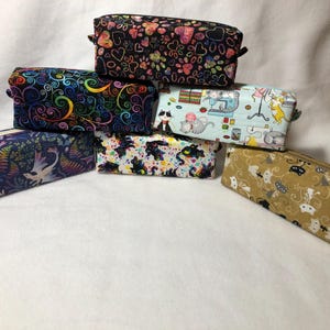 May include: A collection of rectangular fabric pouches with various patterns. Designs include paw prints, hearts, swirls, dragons, cats, and sewing-themed illustrations. Each pouch has a zipper closure and small fabric loops on the sides.