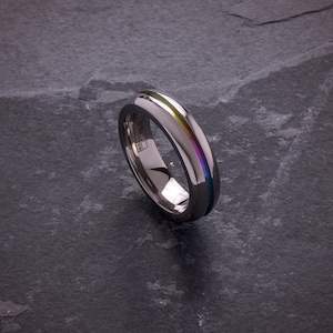 May include: A polished silver ring with a rainbow-colored stripe running down the center. The ring has a smooth, reflective surface and a rounded shape. The rainbow stripe includes colors like yellow, green, blue, and purple.