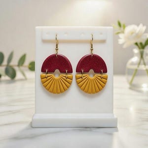 Può includere: Un paio di orecchini fatti a mano. La parte superiore è rosso scuro con glitter, e la parte inferiore presenta un motivo a raggiera giallo senape. Gli orecchini pendono da ganci color oro.