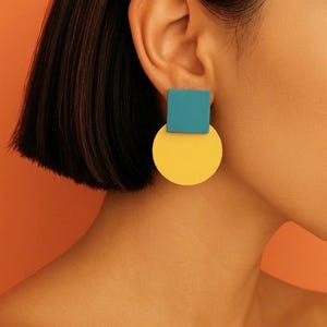 May include: A geometric earring featuring a teal square top piece connected to a larger, solid yellow circular bottom piece. The earring has a smooth, matte finish and a clean, simple design that hangs directly from the earlobe.