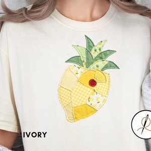 May include: Ivory t-shirt with a patchwork pineapple design. The pineapple features yellow and green sections with a red accent. The word "IVORY" is printed on the shirt. The shirt is a casual, comfortable style.