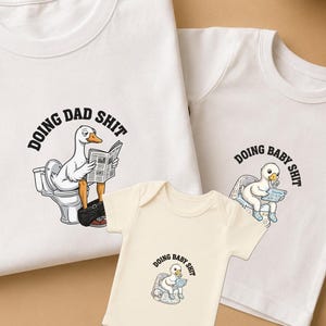 May include: A set of white t-shirts and a onesie with a humorous design. The shirts and onesie feature a cartoon goose on a toilet reading a newspaper, with the phrases "DOING DAD SHIT" and "DOING BABY SHIT".