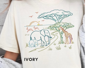 May include: An ivory t-shirt featuring a colorful line art graphic of an African savanna scene. The design includes an elephant with a calf, a giraffe, acacia trees, a sun, and birds. The word IVORY is printed in black below the graphic.