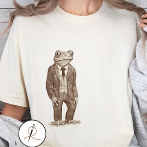Könnte beinhalten: Cremefarbenes T-Shirt mit einer detaillierten Illustration eines Frosches in Anzug und Krawatte. Der Frosch ist im Vintage-Stil dargestellt und trägt einen dreiteiligen Anzug. Das Design ist auf dem Shirt zentriert und bietet eine einzigartige und skurrile Ästhetik.