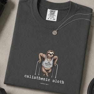 Könnte beinhalten: Dunkelgraues Comfort Colors T-Shirt mit einem Cartoon-Faultier, das Calisthenics macht. Das Faultier trägt ein weißes Tanktop mit dem Wort "SLOTH". Der Text "calisthenic sloth" ist darunter gedruckt.
