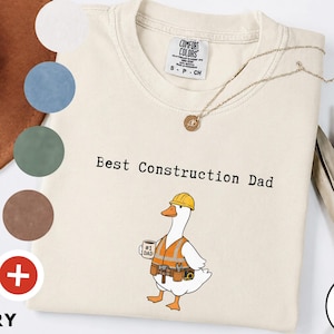 Pode incluir: Camiseta cor marfim com a frase "Best Construction Dad" e um desenho de um ganso com colete e capacete de construção. O ganso segura uma caneca com "#1 Dad". Um colar de ouro e amostras de cores também são visíveis.