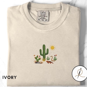 May include: Ivory-colored t-shirt with a crew neck. The shirt features an embroidered desert scene with a green cactus, a yellow sun, and a lizard. The tag reads "COMFORT COLORS" and "XL - TG - EG".