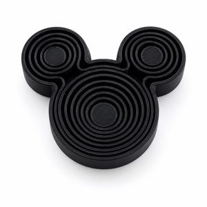 Puede incluir: Un objeto negro tridimensional con forma de cabeza estilizada y dos orejas circulares. La superficie presenta una serie de anillos conc&eacute;ntricos en relieve que crean un patr&oacute;n texturizado y superpuesto en toda la cara y las secciones de las orejas.