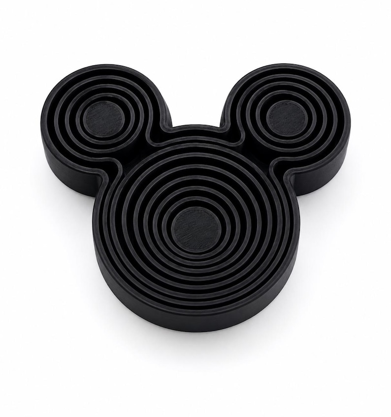 Puede incluir: Un objeto negro tridimensional con forma de cabeza estilizada y dos orejas circulares. La superficie presenta una serie de anillos conc&eacute;ntricos en relieve que crean un patr&oacute;n texturizado y superpuesto en toda la cara y las secciones de las orejas.