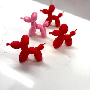 Puede incluir: Se muestran cuatro figuras de perros globo. Tres son rojos y uno es rosa. Las esculturas de perros están sobre una superficie blanca. Los perros globo son un objeto decorativo caprichoso.