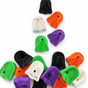 May include: A collection of ghost-shaped cookie cutters in black, white, orange, purple, and green. Each cutter features a layered, ridged design with a hollow center, arranged in a scattered pattern against a plain white background.