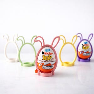 May include: A selection of Kinder Joy eggs displayed in bunny-shaped holders. The holders are in pastel shades of pink, yellow, green, white, and purple. The Kinder Joy eggs have a white and orange shell with the words "Kinder Joy" printed on them.