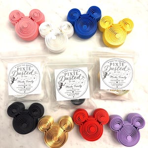 May include: A collection of eight Mickey Mouse-shaped plastic icons in pink, white, blue, yellow, black, gold, red, and purple. Three icons are inside clear plastic bags with labels reading You've Been Pixie Dusted by the Meeks Family Stateroom 7186.