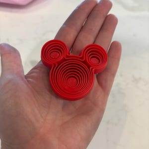 Puede incluir: Un objeto rojo brillante en tres dimensiones con forma de cabeza de rat&oacute;n estilizada. El dise&ntilde;o presenta crestas circulares conc&eacute;ntricas que crean un efecto de capas escalonadas dentro de la cabeza principal y las dos orejas peque&ntilde;as.