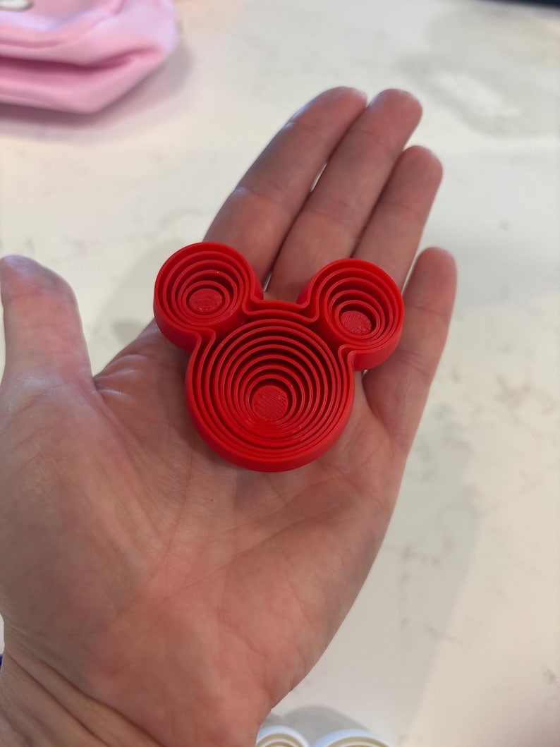 Puede incluir: Un objeto rojo brillante en tres dimensiones con forma de cabeza de rat&oacute;n estilizada. El dise&ntilde;o presenta crestas circulares conc&eacute;ntricas que crean un efecto de capas escalonadas dentro de la cabeza principal y las dos orejas peque&ntilde;as.