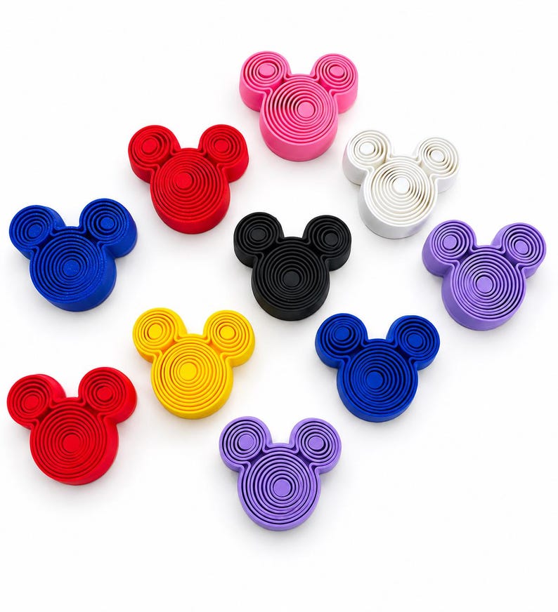 Puede incluir: Una colecci&oacute;n de once cepillos de limpieza facial de silicona con forma de cabeza de rat&oacute;n en varios colores, incluyendo rosa, rojo, blanco, negro, morado, amarillo y azul. Cada cepillo presenta una serie de surcos circulares conc&eacute;ntricos.
