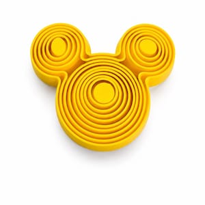 Puede incluir: Un objeto tridimensional de color amarillo brillante con forma de cabeza estilizada y dos orejas circulares. La superficie presenta crestas circulares conc&eacute;ntricas en relieve que crean un efecto topogr&aacute;fico en capas sobre toda la forma del objeto.