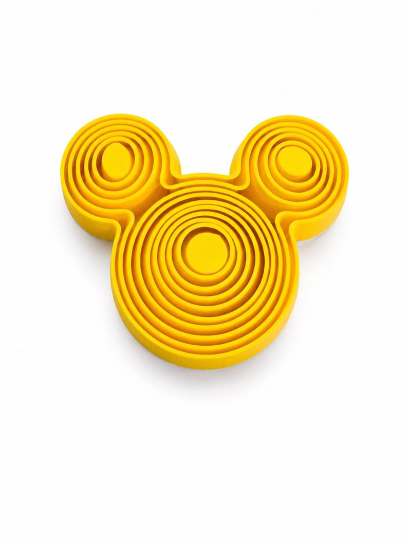 Puede incluir: Un objeto tridimensional de color amarillo brillante con forma de cabeza estilizada y dos orejas circulares. La superficie presenta crestas circulares conc&eacute;ntricas en relieve que crean un efecto topogr&aacute;fico en capas sobre toda la forma del objeto.