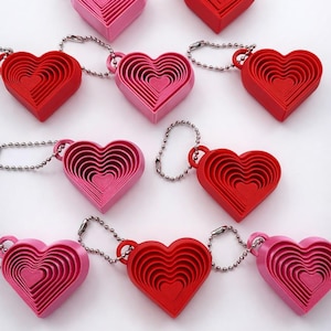Könnte beinhalten: Eine Sammlung herzförmiger Schlüsselanhänger in Rot und Rosa. Jeder Schlüsselanhänger hat ein verschachteltes Design mit mehreren Herzschichten. Die Schlüsselanhänger sind an silbernen Kugelkettchen befestigt, was eine ansprechende Anordnung ergibt.