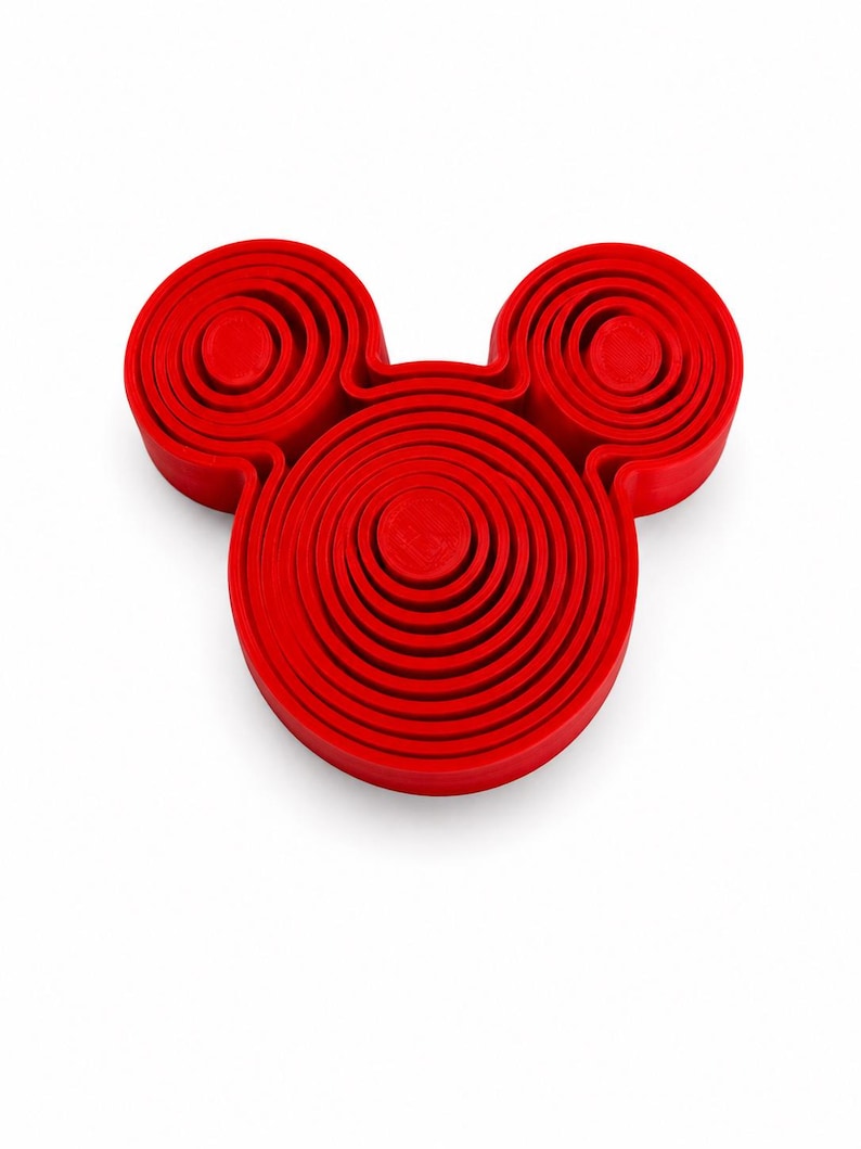 Puede incluir: Un objeto de color rojo brillante con base circular que presenta un anillo central grande y dos anillos m&aacute;s peque&ntilde;os colocados encima, creando una silueta que recuerda a una cabeza con dos orejas. La superficie tiene m&uacute;ltiples crestas circulares conc&eacute;ntricas.