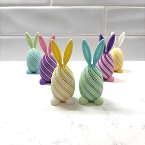 Puede incluir: Un conjunto de figuras de conejitos de Pascua coloridos. Cada conejito tiene forma de huevo con patrones de rayas en varios colores pastel, incluyendo azul, verde, morado y amarillo. Los conejitos tienen orejas erguidas y pequeños pies blancos.