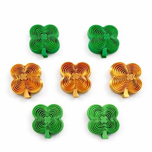 Op de afbeelding: Zeven klavervormige objecten in groen- en goudtinten zijn gerangschikt op een witte achtergrond. De objecten hebben een gelaagd ontwerp, waardoor een driedimensionaal effect ontstaat. De groene klavers hebben een steel, de gouden niet.