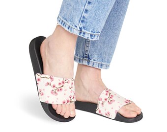 May include: Black slide sandals with a white upper featuring a pink cherry blossom design. The sandals are worn with blue jeans. The floral pattern includes pink flowers and small pink petals.