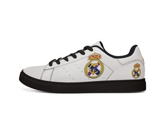 Puede incluir: Zapatilla blanca con cordones negros y suela negra. Presenta el escudo del Real Madrid en dorado, azul y amarillo en el lateral. Un escudo más pequeño en el talón. Diseño clásico y de perfil bajo.