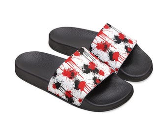May include: A pair of black slide sandals with a white strap featuring a pattern of red and black paint splatters. The sandals have a textured footbed and a solid black sole, perfect for casual wear.