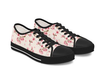 May include: A pair of canvas sneakers with a floral pattern. The shoes have a cream-colored background with pink cherry blossom branches. The toe caps and soles are black, with black laces and a white stripe on the sole.