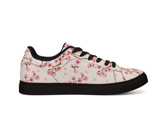 May include: White sneakers with a black sole and laces. The shoes are decorated with a pink cherry blossom pattern. The design features pink flowers and branches on a white background, creating a floral theme.