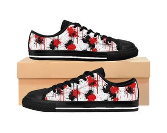 May include: A pair of canvas sneakers with a black toe cap and sole. The white canvas uppers are decorated with a pattern of red and black paint splatters. The shoes are displayed on a brown cardboard box.
