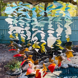 May include: A colorful wind chime made of various shapes of sea glass. The sea glass pieces are in shades of blue, white, yellow, orange, and red, suspended from a natural wooden branch. The wind chime hangs outdoors in front of a blue fence.