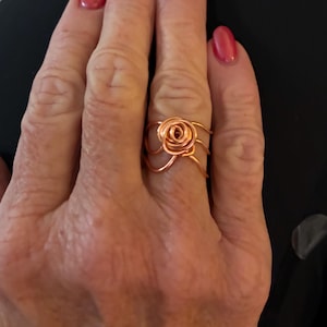 May include: A copper-colored wire ring featuring a rose design. The ring's floral element is meticulously crafted with delicate wire, creating a three-dimensional rose. The ring is a unique piece of jewelry.