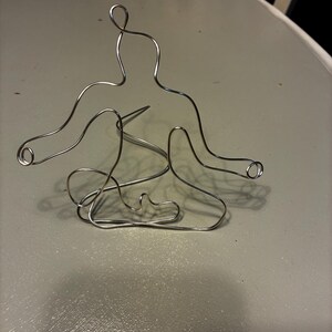 May include: A silver wire sculpture of a person in a seated yoga pose. The sculpture is made of a single piece of wire, with rounded ends on the arms. The figure is sitting on a light gray surface.