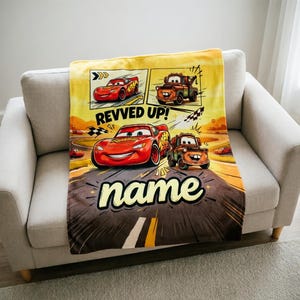 May include: A yellow, brown, and red Cars-themed blanket featuring Lightning McQueen and Mater. The blanket has a comic book style design with the words "Revved Up!" and "name" in a bold font.