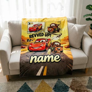 May include: A yellow and brown blanket featuring cartoon cars. The blanket has a road design with the text "REVVED UP!" and "name". The blanket is draped over a gray sofa.