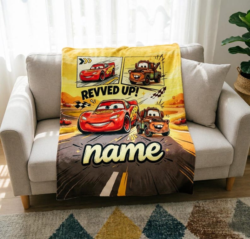 May include: A yellow and brown blanket featuring cartoon cars. The blanket has a road design with the text "REVVED UP!" and "name". The blanket is draped over a gray sofa.