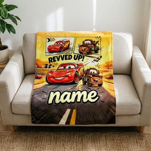 May include: A plush blanket featuring Lightning McQueen and Mater from the Cars movie. The blanket has a yellow and brown background with the words "REVVED UP!" and "name" in a playful font. The blanket is draped over a beige sofa.