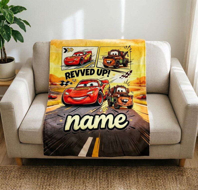 May include: A plush blanket featuring Lightning McQueen and Mater from the Cars movie. The blanket has a yellow and brown background with the words "REVVED UP!" and "name" in a playful font. The blanket is draped over a beige sofa.